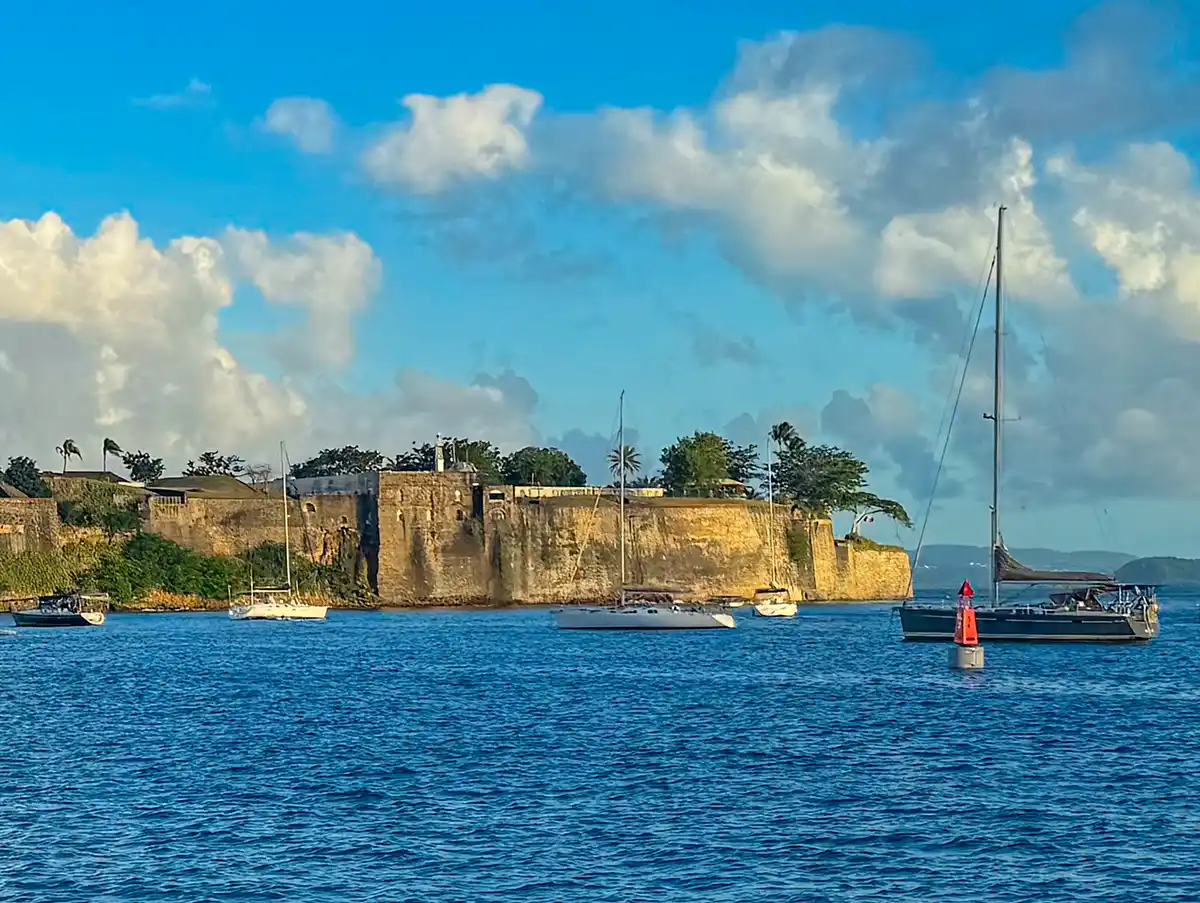 Sehenswuerdigkeiten-Martinique-Fort-Saint-Louis Blaues Wasser mit Segelbooten, die in der Nähe eines alten Steinforts unter einem teilweise bewölkten Himmel vor Anker liegen.