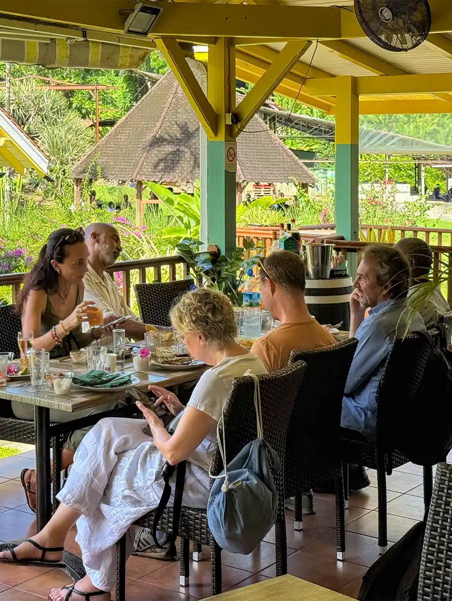 Sehenswuerdigkeiten-Martinique-Restaurant-Bananenmuseum Sechs Personen sitzen um einen Tisch in einem offenen Restaurant, unterhalten sich und entspannen sich in einer üppigen, tropischen Umgebung.
