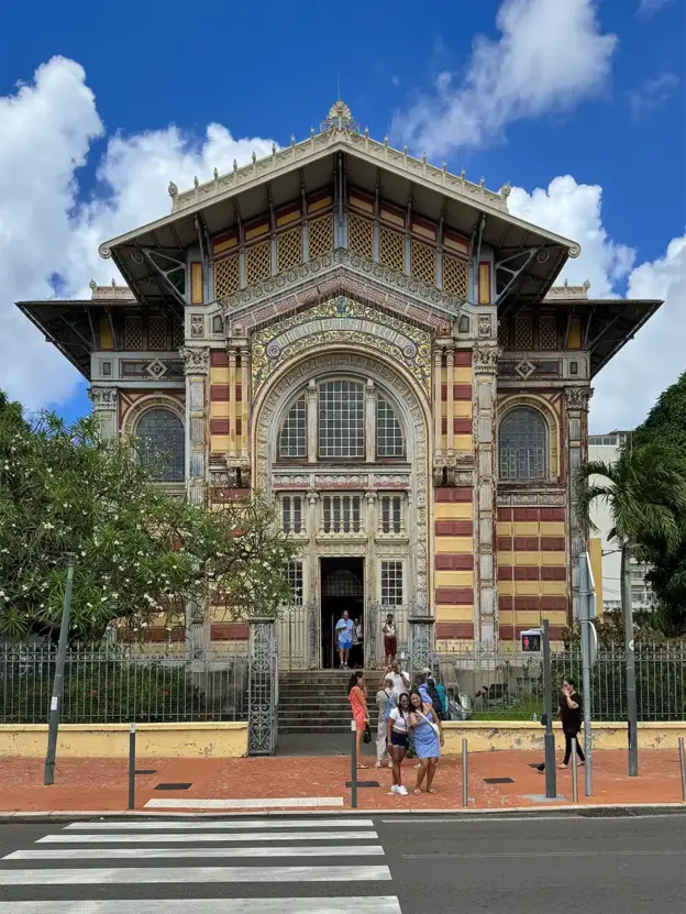 Ein farbenfrohes, verziertes Gebäude mit gewölbten Fenstern und davor stehenden Menschen, gesehen über einen Zebrastreifen.
