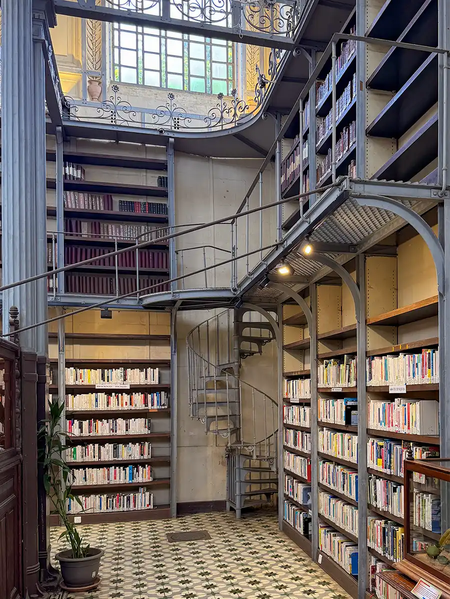 Sehenswurdigkeiten-Martinique-Bibliothek-Schoelcher-innen Eine Bibliothek mit hohen Bücherregalen, einer Wendeltreppe und dekorativen Metallgeländern unter einem großen Fenster.