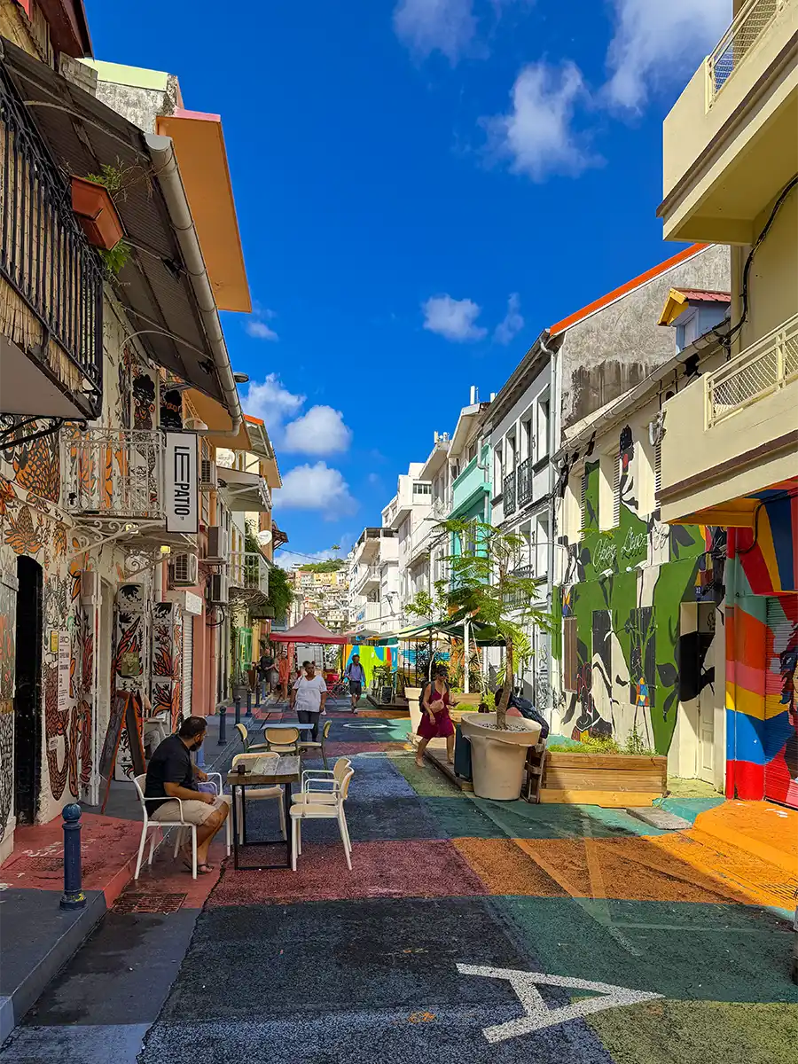 Sehenswurdigkeiten-Martinique-Fort-de-France-Rue-coloree Eine farbenfrohe Straße mit Wandmalereien, Sitzgelegenheiten im Freien, Menschen, die spazieren gehen, und strahlend blauem Himmel über der Stadt.