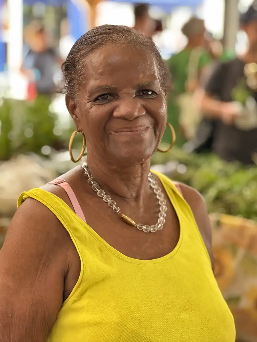 Sehenswurdigkeiten-Martinique-Markt-Kraeuterfrau-Jeronie Lächelnde ältere Frau in einem gelben Tank-Top und Schmuck auf einem Markt im Freien.