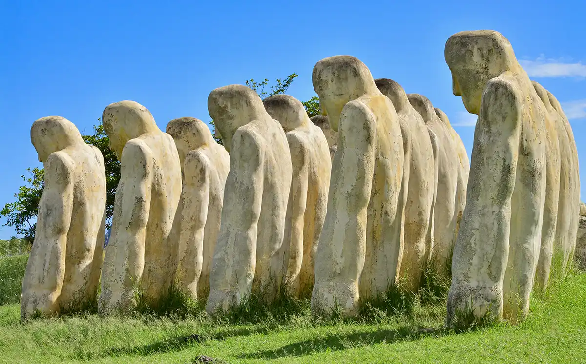 Sehenswurdigkeiten-Martinique-Sklavenmonument Eine Reihe großer menschlicher Steinfiguren steht mit gesenkten Köpfen auf grasbewachsenem Boden unter blauem Himmel.