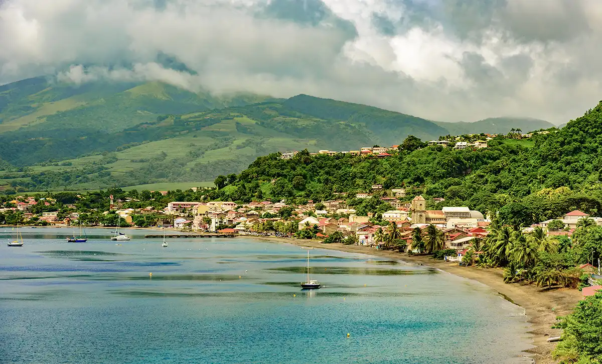 Sehenswurdigkeiten-Martinique-St-Pierre Küstenstadt mit bunten Gebäuden, Sandstrand, Booten, üppig grünen Hügeln und Bergen unter bewölktem Himmel.