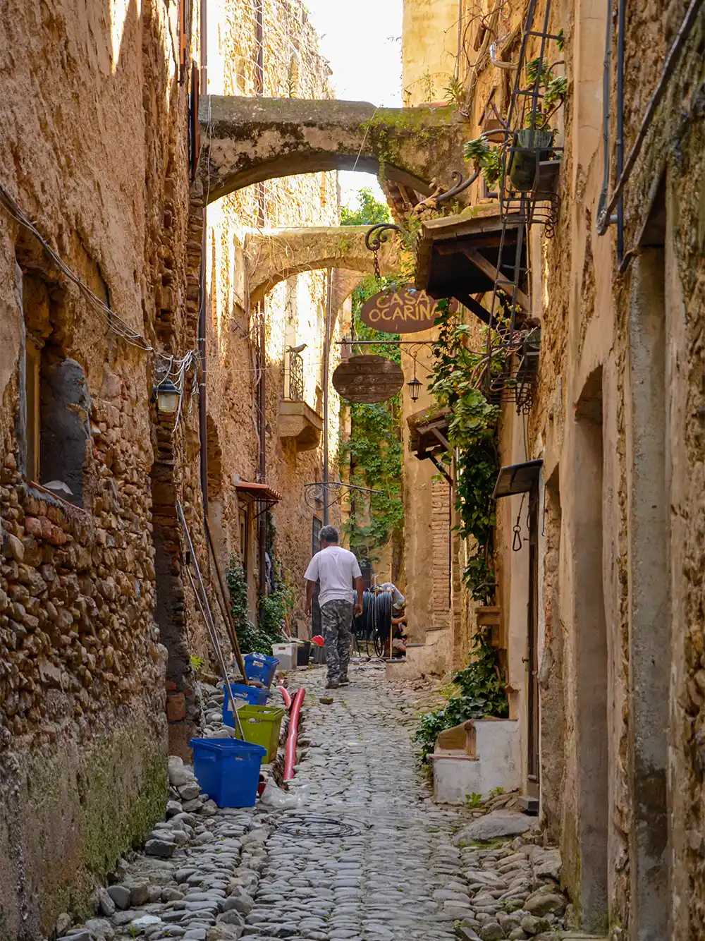 Eine schmale Kopfsteinpflastergasse mit Steinmauern, hängenden Schildern, Topfpflanzen und Menschen, die weggehen.