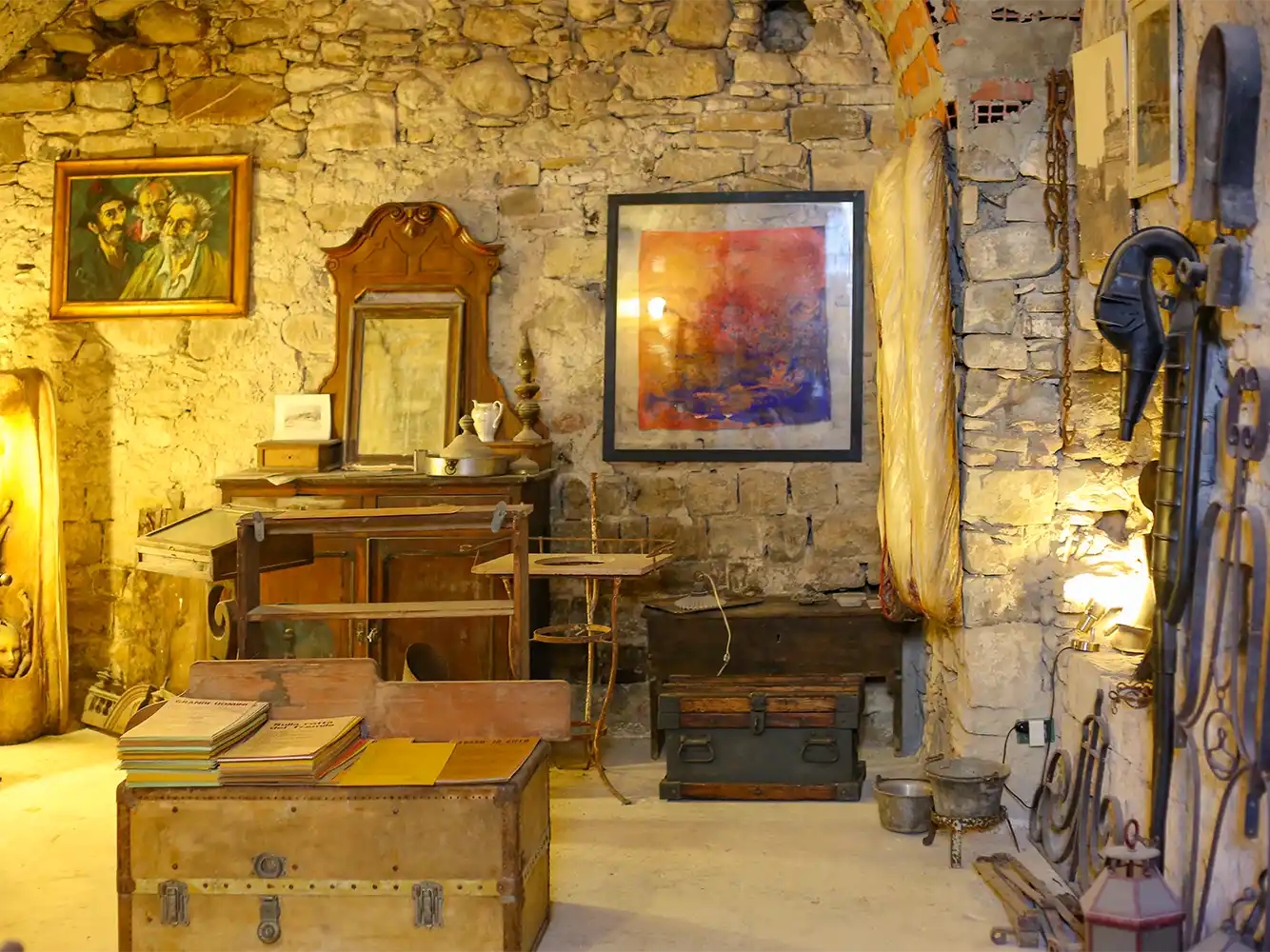 Ein rustikaler Steinsaal mit alten Möbeln, Gemälden, Koffern und verschiedenen antiken Gegenständen, die ausgestellt sind.