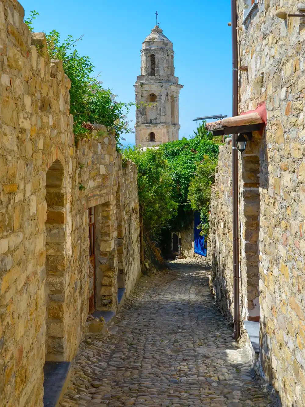 Enge Kopfsteinpflastergasse mit Steinhäusern und einem Glockenturm im Hintergrund an einem sonnigen Tag.