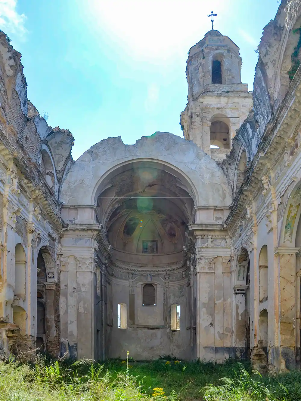 Ruinen einer alten, dachlosen Kirche mit überwuchertem Gras und Sonnenlicht, das durch die offene Decke fällt.
