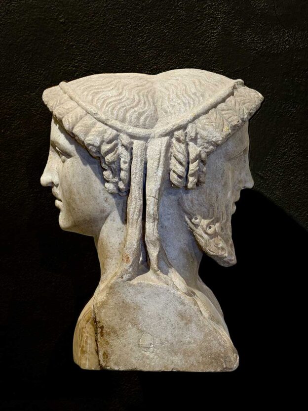 Antike doppelgesichtige Marmorbüste vor dunklem Hintergrund, die zwei Köpfe zeigt, die am Rücken verbunden sind.