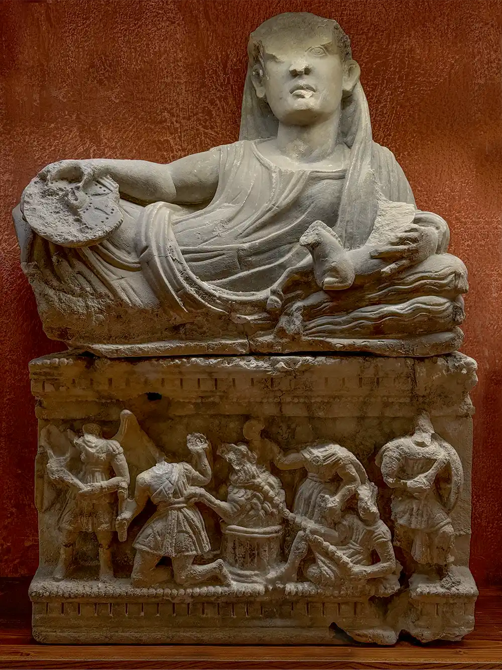 Antike Marmorstatue einer liegenden Figur über detaillierten Schnitzereien von Soldaten, vor einem rötlichen Hintergrund.