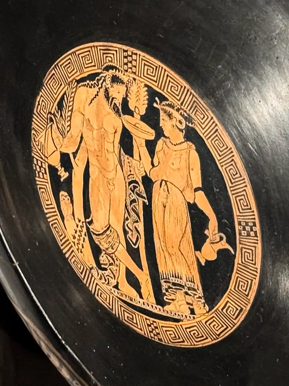 Antike griechische Keramik mit zwei etruskischen Frauenfiguren in traditioneller Tracht und einem geometrischen Rand auf schwarzem Hintergrund.