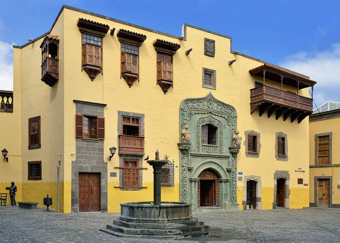Historisches gelbes Gebäude mit verschnörkeltem Steintor, Holzbalkonen und einem Springbrunnen auf einem mit Kopfstein gepflasterten Platz.