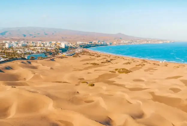 Goldene Sanddünen erstrecken sich entlang der Küste, mit einer Stadt und Bergen im Hintergrund unter einem klaren Himmel.