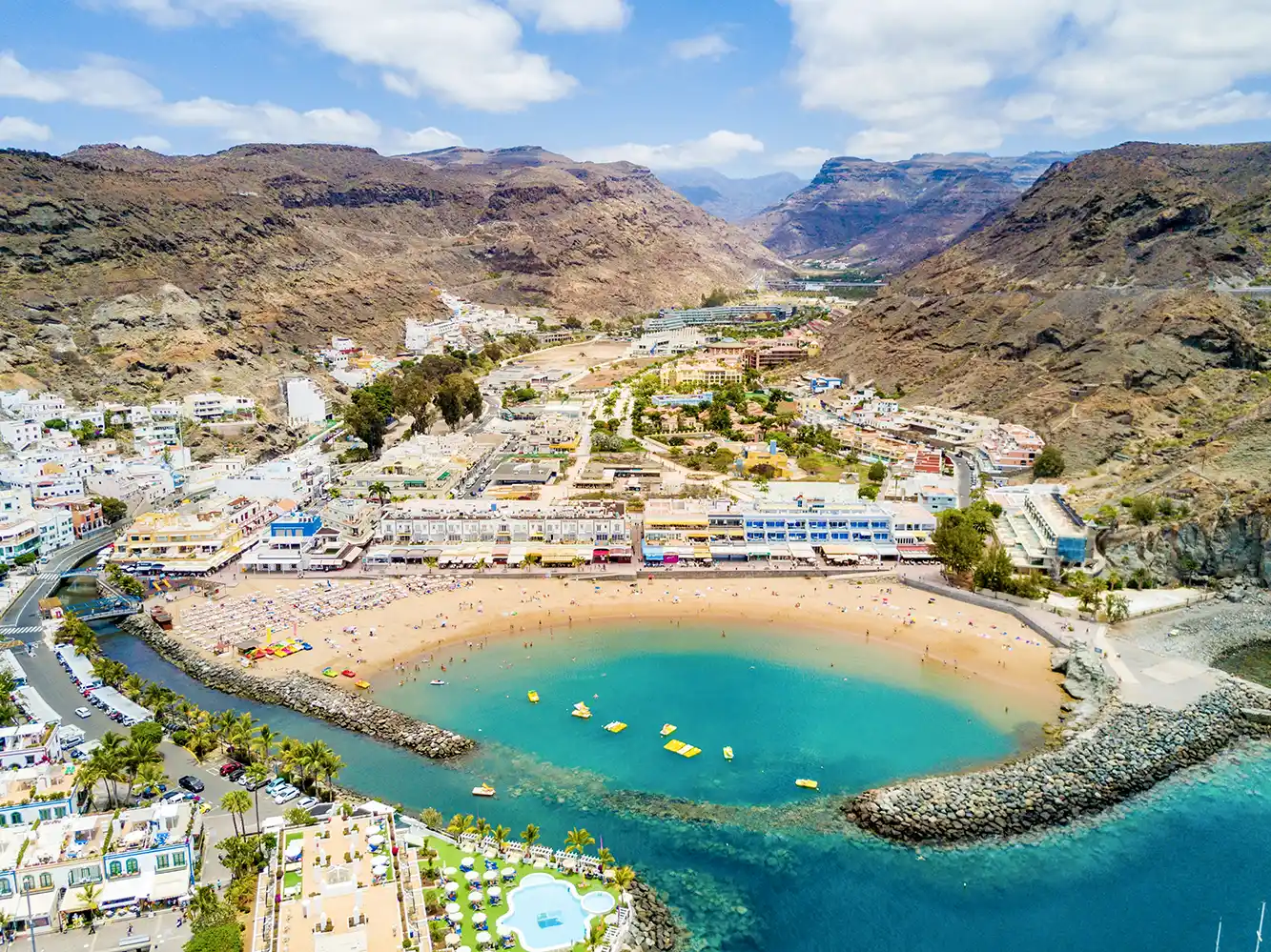 Luftaufnahme eines Sandstrandes mit türkisfarbenem Wasser, umgeben von Bergen und Gebäuden.