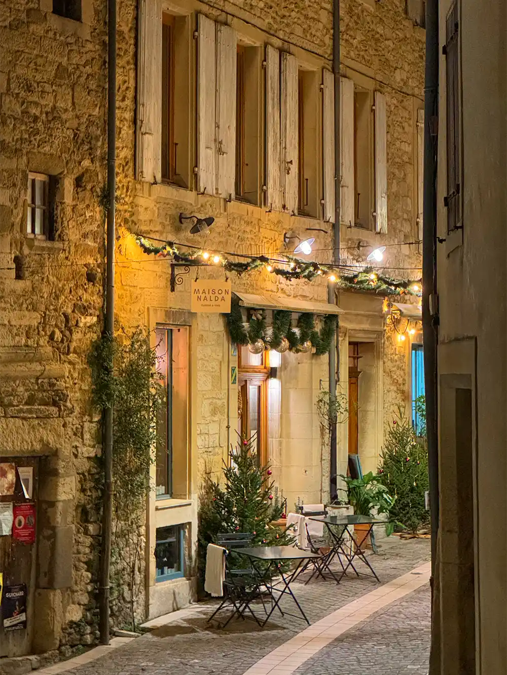 Grignan-Gasse-Restaurant Außencafé mit leeren Tischen in einer schmalen, gepflasterten Straße neben einem warm beleuchteten, mit Girlanden geschmückten Steingebäude.