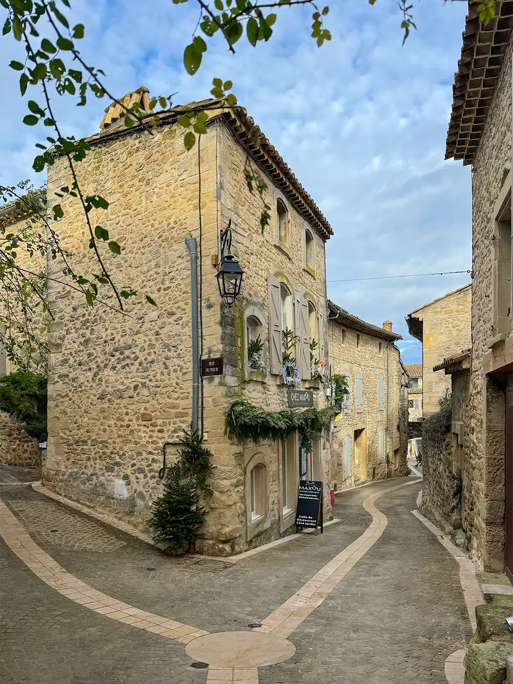 Grignan-Gassen Eine schmale Kopfsteinpflasterstraße schlängelt sich zwischen steinernen Gebäuden in einem malerischen europäischen Dorf unter einem teilweise bewölkten Himmel.