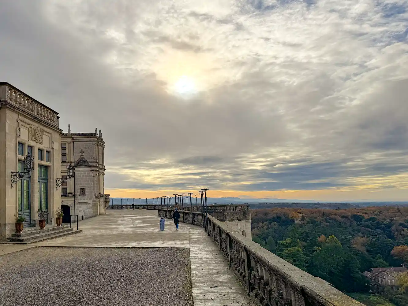 Grignan-Provence-Schloss-Landschaft Ein bewölkter Himmel über einer Schlossterrasse mit zwei Spaziergängern und einer Waldlandschaft in der Ferne.
