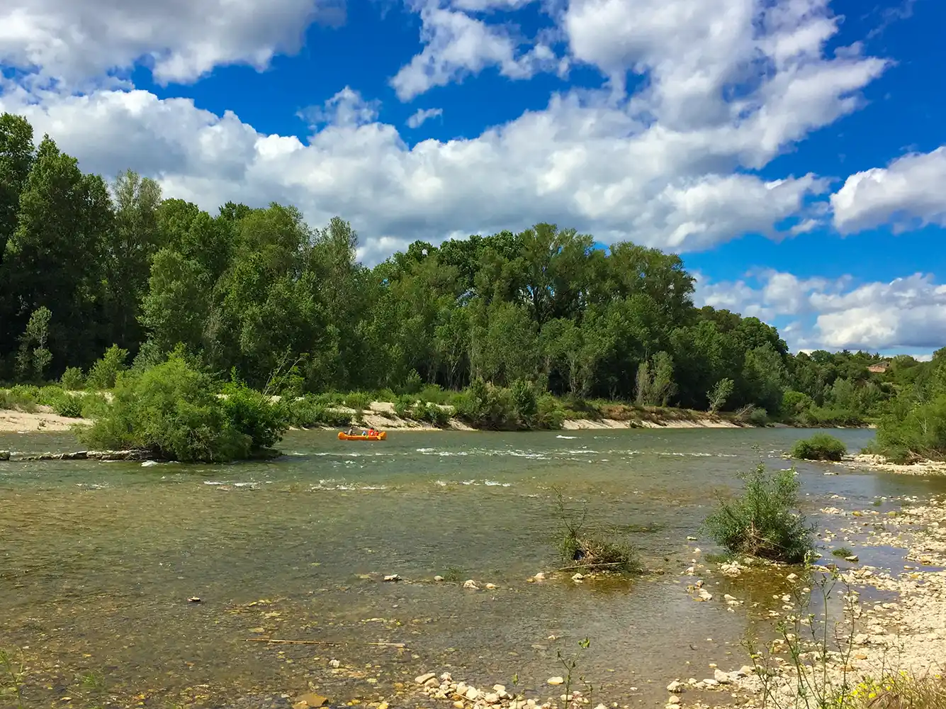 Ein breiter, flacher Fluss fließt an grünen Bäumen vorbei unter einem blauen Himmel mit weißen Wolken; ein Floß ist auf dem Wasser.