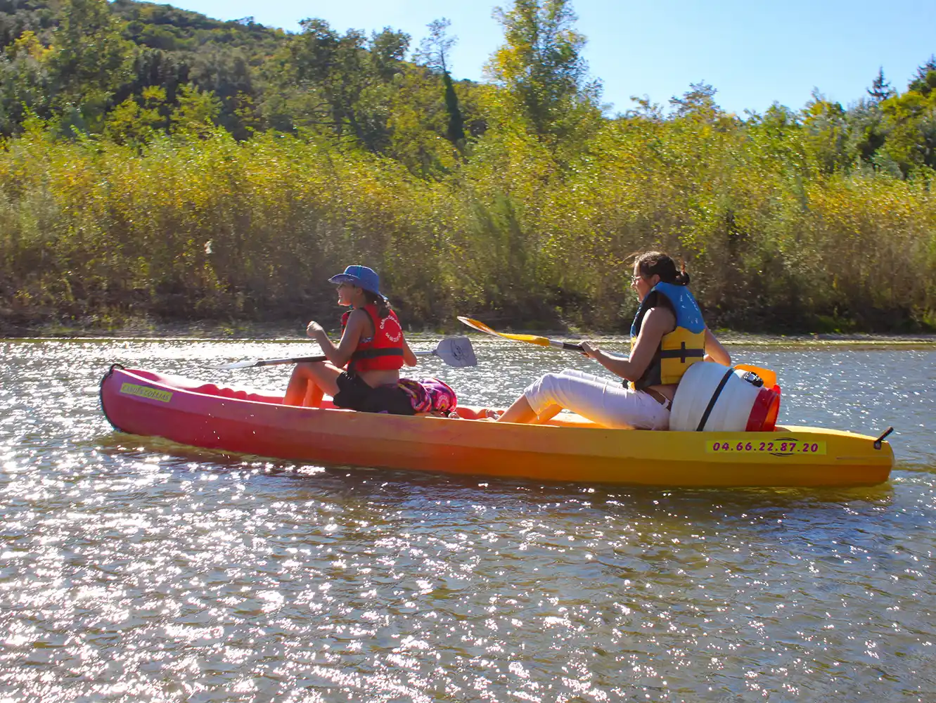 Zwei Personen fahren Kajak auf einem sonnenbeschienenen Fluss, umgeben von Grün und Bäumen im Hintergrund.