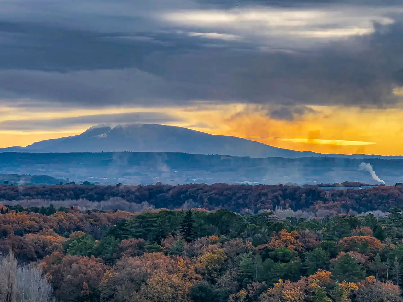 Mont-Ventoux Sonnenuntergang über einem Wald mit aufsteigendem Rauch und einem entfernten Berg unter einem dramatischen, bewölkten Himmel.