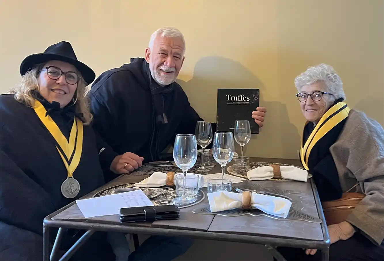 Richerenches-Interview-Elisabeth-Fontannaz Drei lächelnde Menschen mit Medaillen sitzen an einem Tisch mit leeren Gläsern und Servietten und halten eine Truffes-Karte in der Hand.