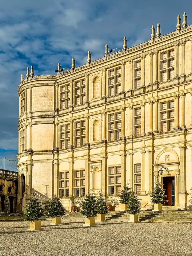 Ein großes Steingebäude mit vielen Fenstern und kleinen Topfbäumen draußen unter einem teilweise bewölkten Himmel.