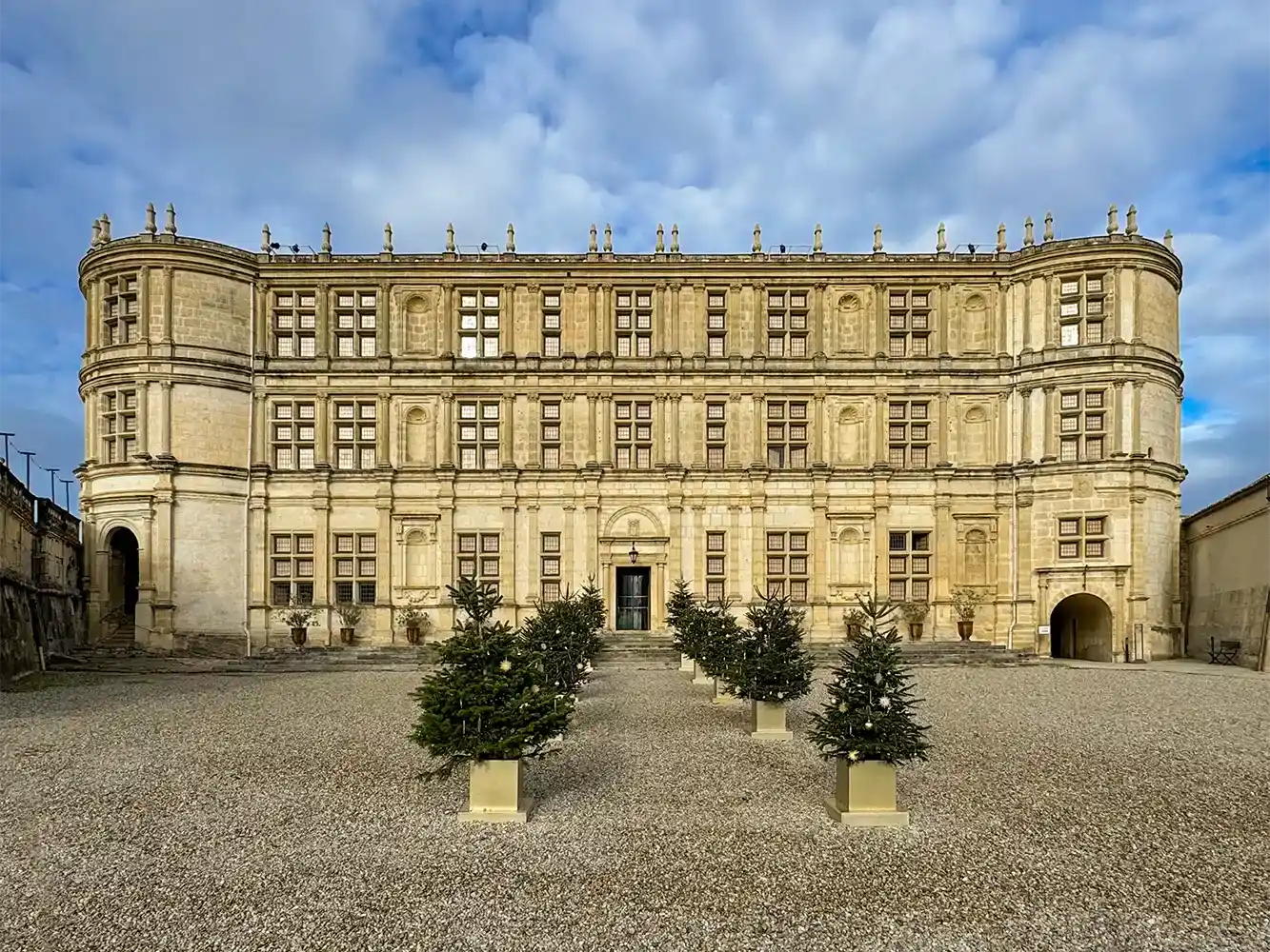 Ein prächtiges historisches Gebäude mit geschmückten Weihnachtsbäumen in Pflanzkübeln, die auf einem Kiesplatz aufgestellt sind.