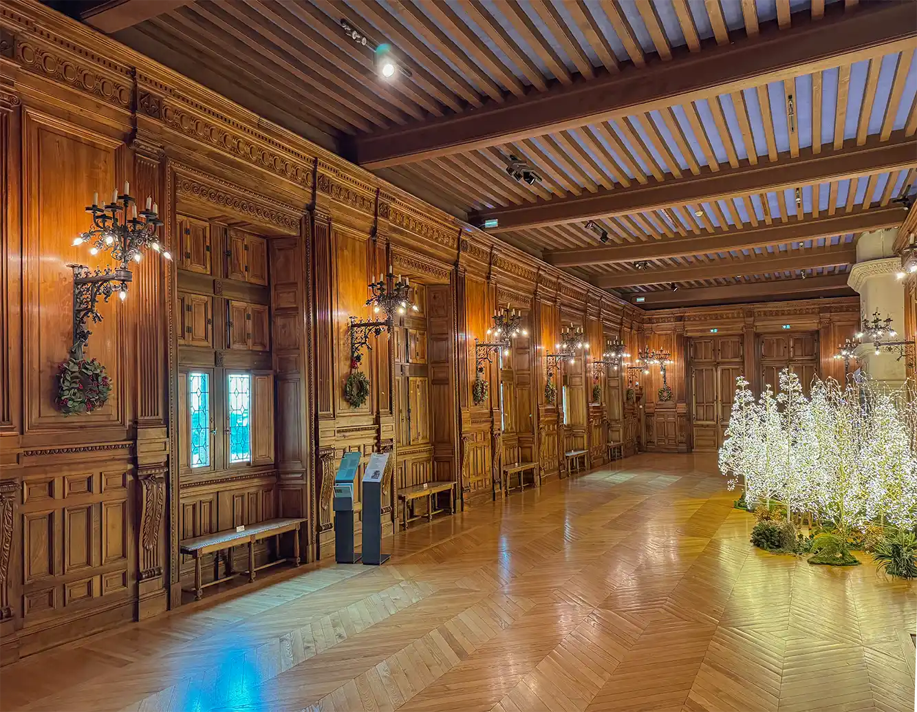 Schloss-Grignan-Galerie-1495 Verzierte holzgetäfelte Halle mit Bänken, Kronleuchtern und beleuchtetem Baumschmuck auf der rechten Seite.