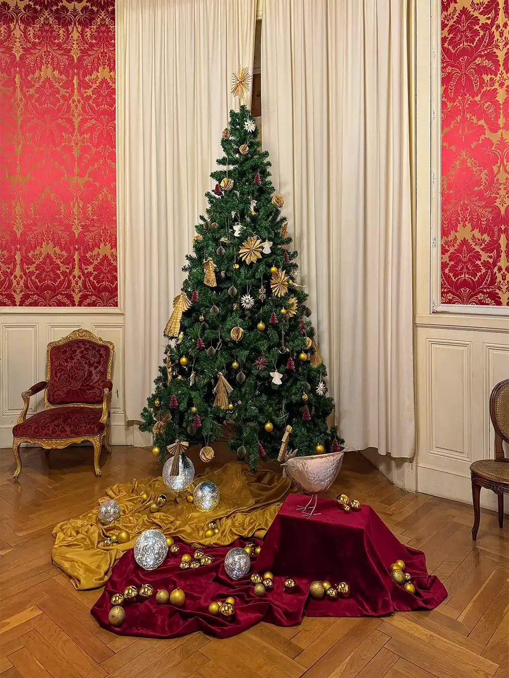Schloss-Grignan-Kabinett-Madame-Grignan Ein geschmückter Weihnachtsbaum mit goldenen Ornamenten und drapiertem Stoff in einem eleganten Raum mit roter Tapete.