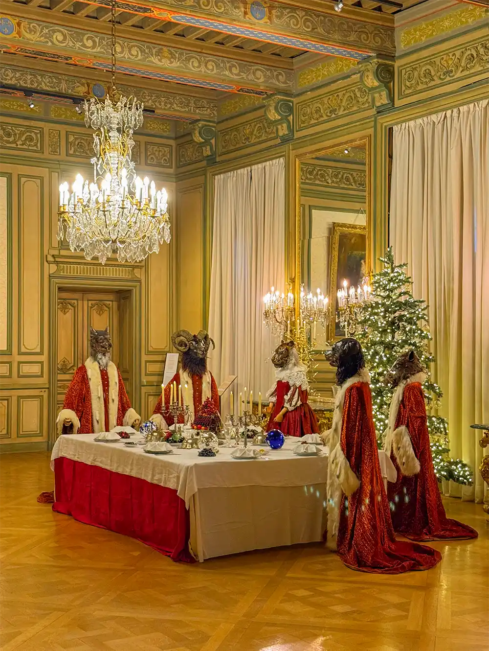 Schloss-Grignan-Koenigssaal Fünf Figuren mit Tiermasken und königlichen Gewändern stehen um einen festlichen, mit Kerzen beleuchteten Esstisch in einem geschmückten Raum.
