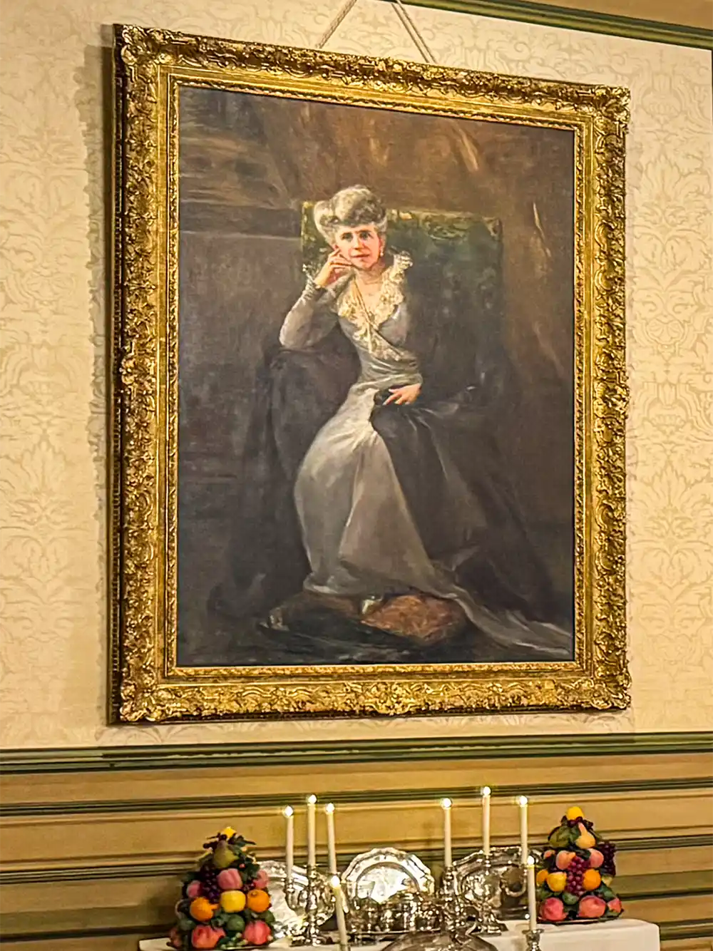 Schloss-Grignan-Marie-Fontaine Ein gerahmtes Porträt einer sitzenden Frau in formeller Kleidung hängt über einem mit Kerzen beleuchteten Tisch mit Obstauslagen.