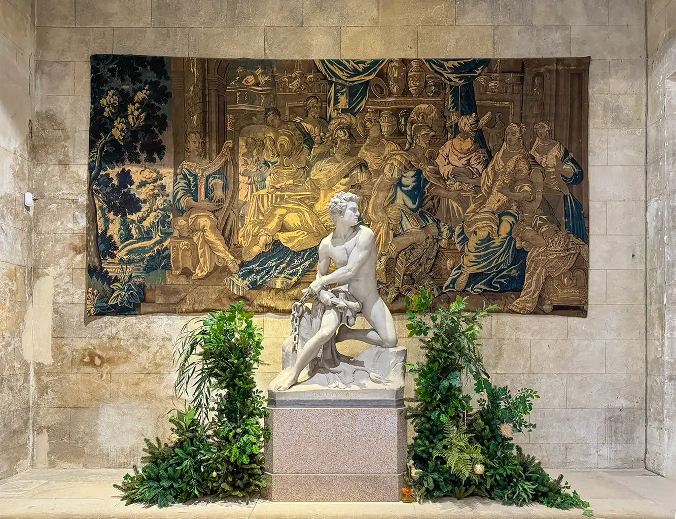 Schloss-Grignan-Wandteppich-Eingangshalle Marmorstatue, umgeben von Pflanzen, vor einem großen, detaillierten Wandteppich an einer Steinwand.