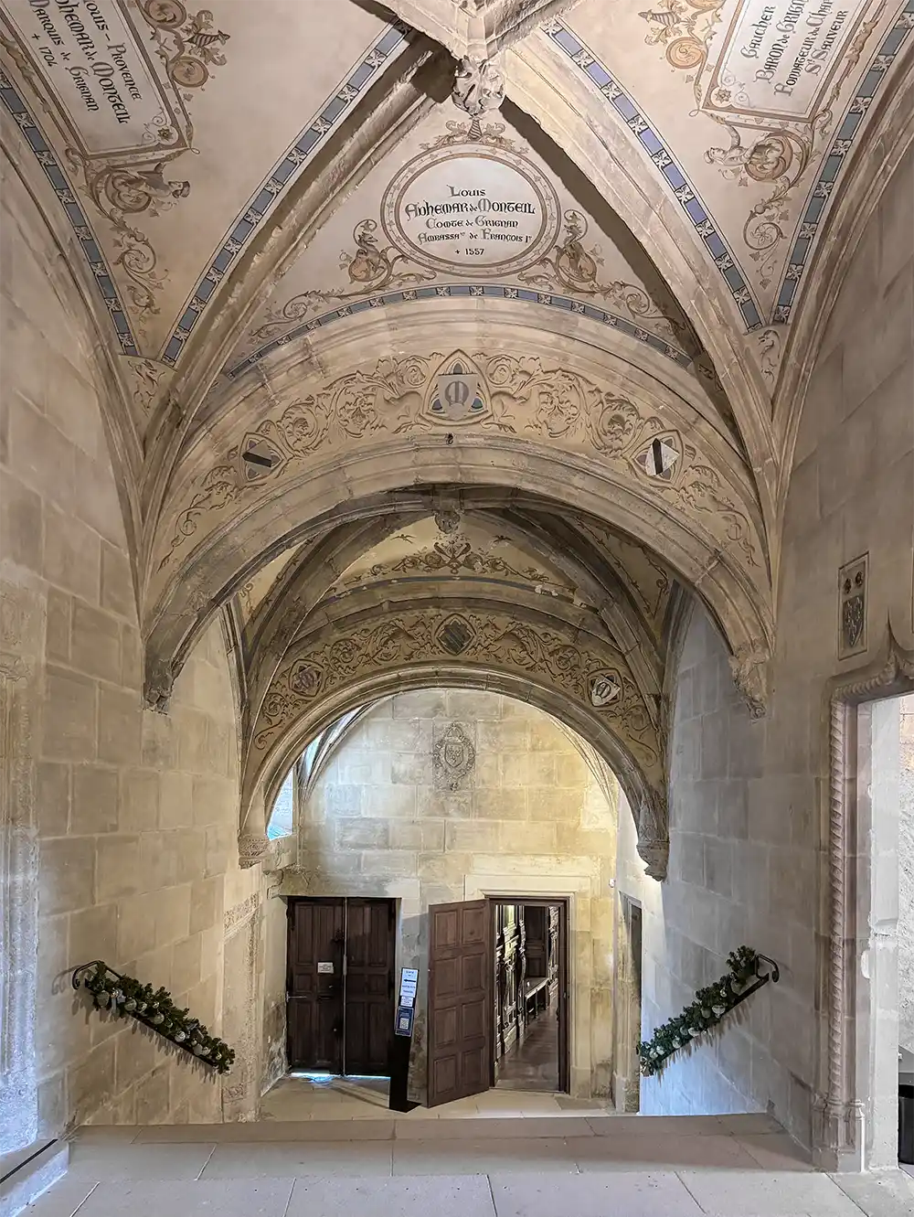 Schloss-Grignan-Wappen Steintreppe mit verzierter Gewölbedecke und offenen Holztüren am unteren Ende, dekoriert mit Grünpflanzen.