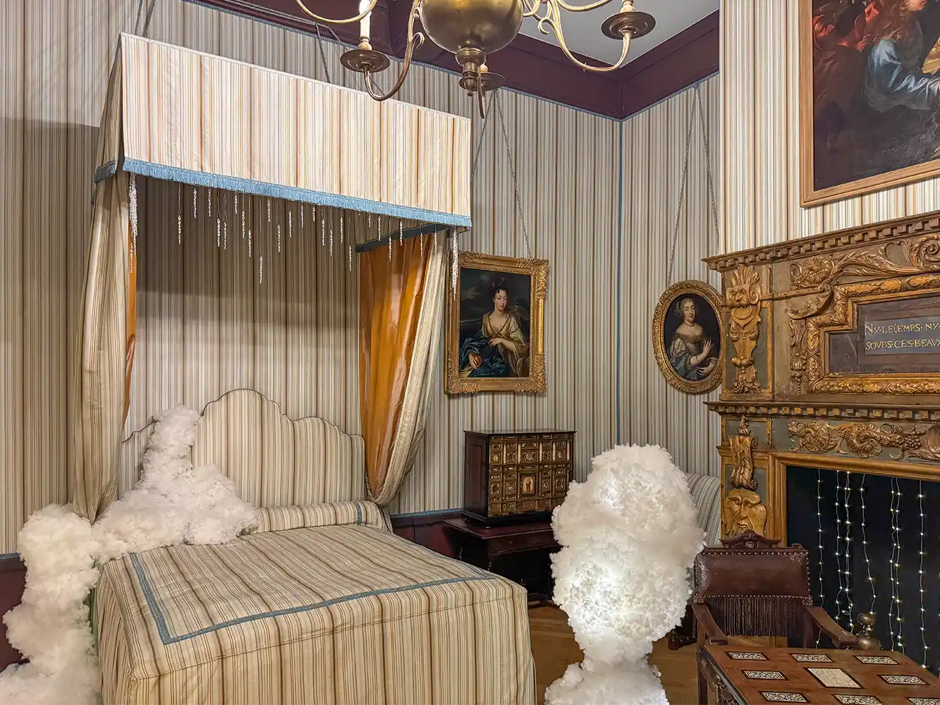 Schloss-Grignan-Zimmer-Madame-Grignan Ein Vintage-Schlafzimmer mit Himmelbett, verschnörkeltem Kamin und gerahmten Porträts an gestreiften Wänden.