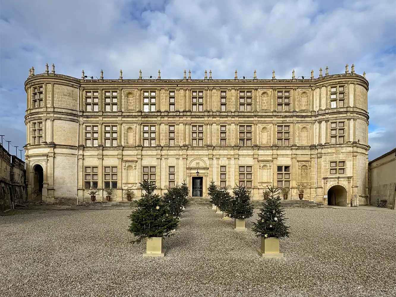 Schloss-Grignan Ein prächtiges historisches Gebäude mit geschmückten Weihnachtsbäumen in Pflanzkübeln, die auf einem Kiesplatz aufgestellt sind.