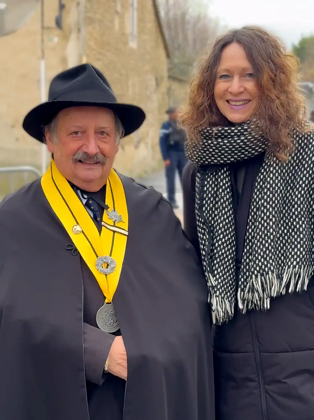 Trueffelmarkt-Richerenches-Bruderschaft Ein lächelnder Mann mit schwarzem Hut und zeremoniellem Umhang steht neben einer Frau in schwarzem Mantel und Schal im Freien.