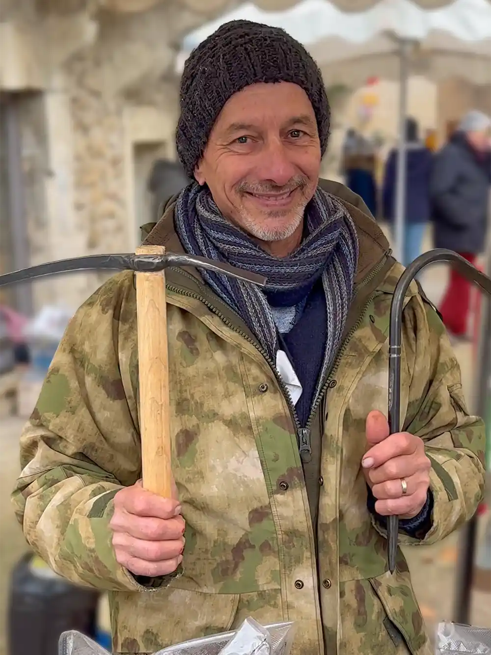 Trueffelmarkt-Richerenches-Christian-Trueffel-Werkzeug Lächelnder Mann mit Mütze und Tarnjacke, der auf dem Trüffelmarkt eine Spitzhacke und eine Art Sichel hält.