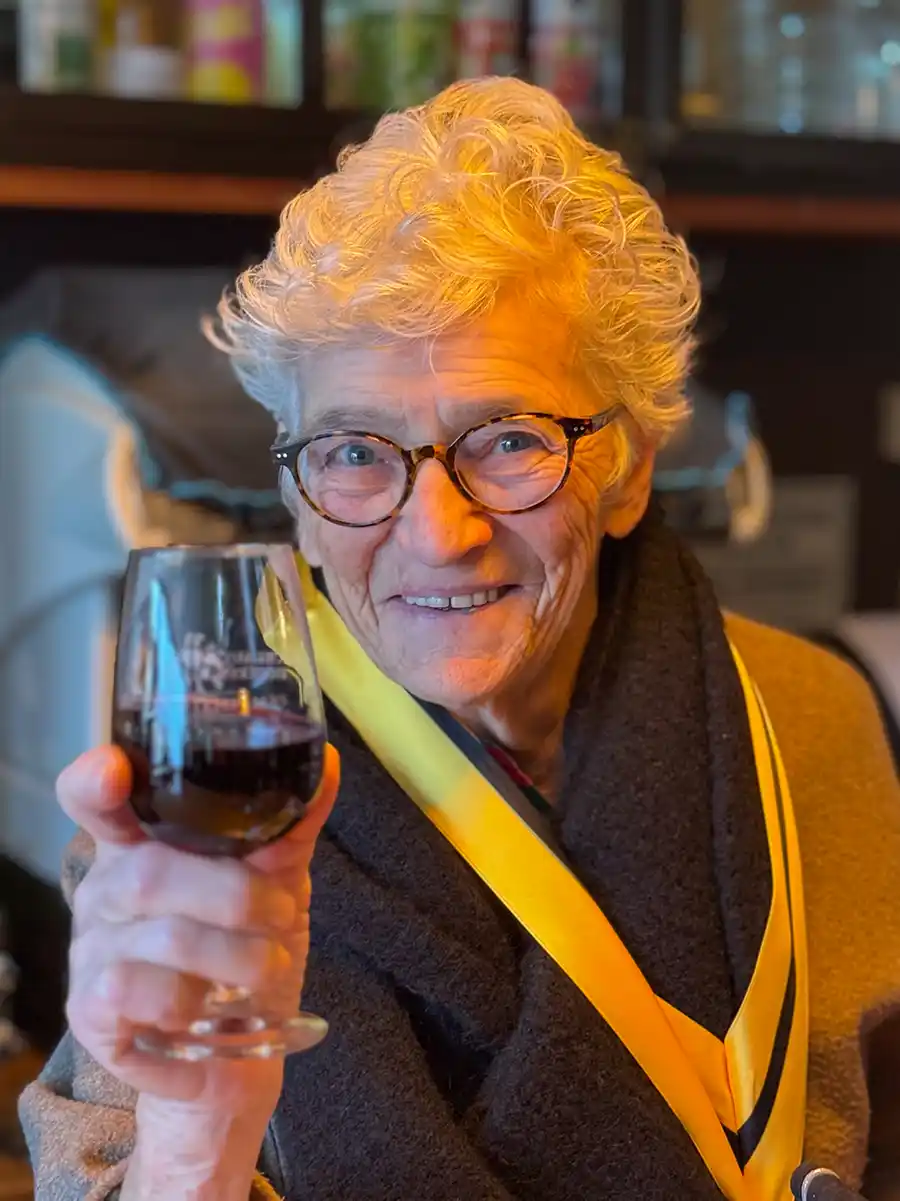 Trueffelmarkt-Richerenches-Elisabeth-Fontannaz Ältere Person mit lockigem Haar und Brille, die lächelnd ein Glas Rotwein hochhält.