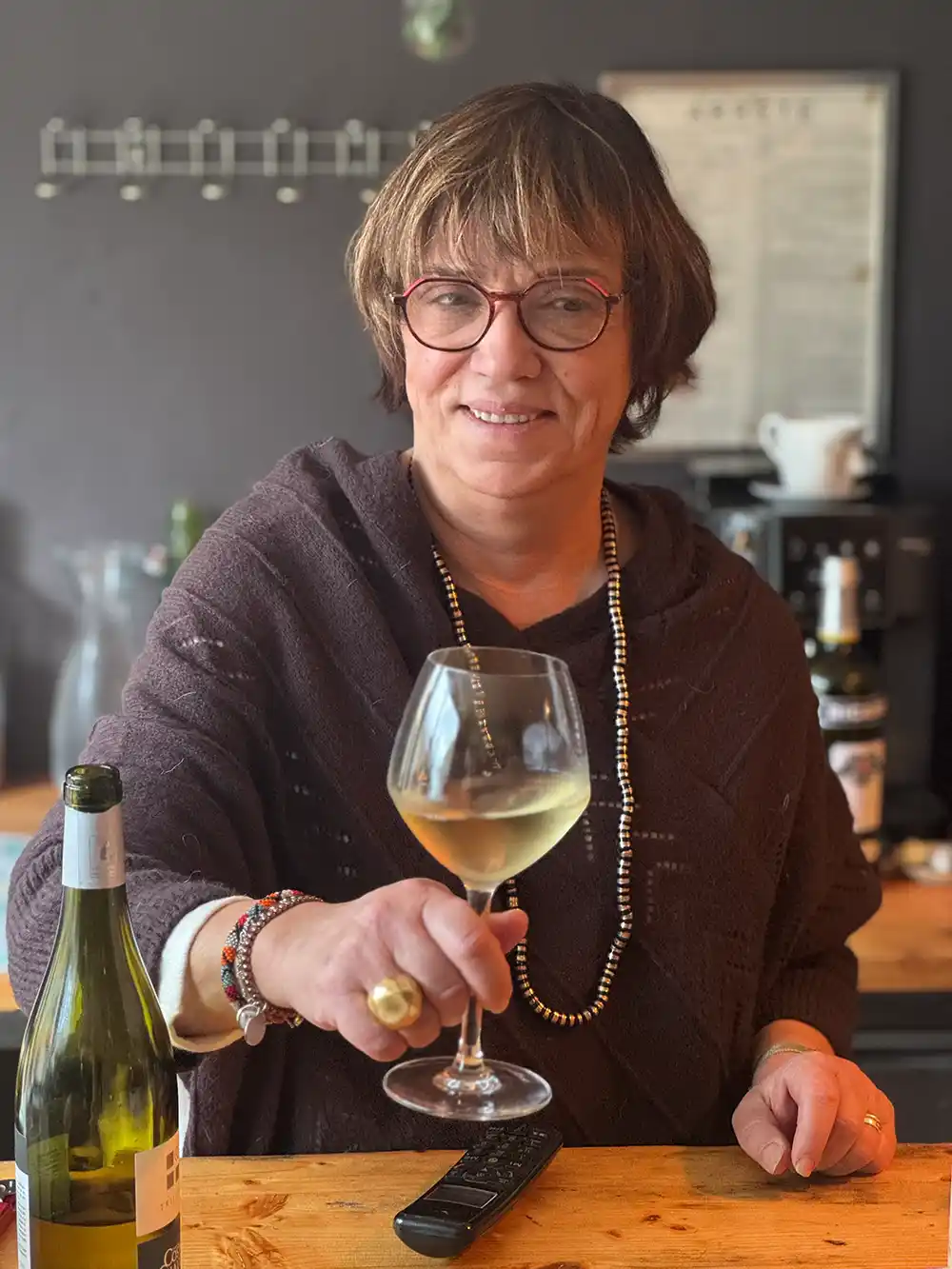 Trueffelmarkt-Richerenches-Sandrine Eine Frau mit kurzen Haaren und Brille lächelt und hält ein Glas Weißwein an einem Holztresen in einem Haus.