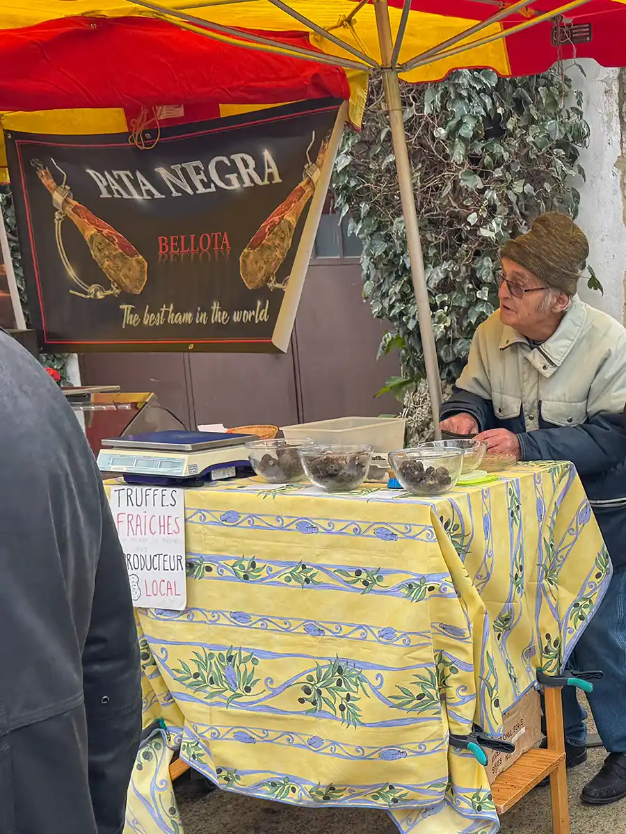 Trueffelmarkt-Richerenches-Schinken Ein Mann sitzt an einem Marktstand, der unter einem rot-gelben Baldachin frische Trüffel verkauft.