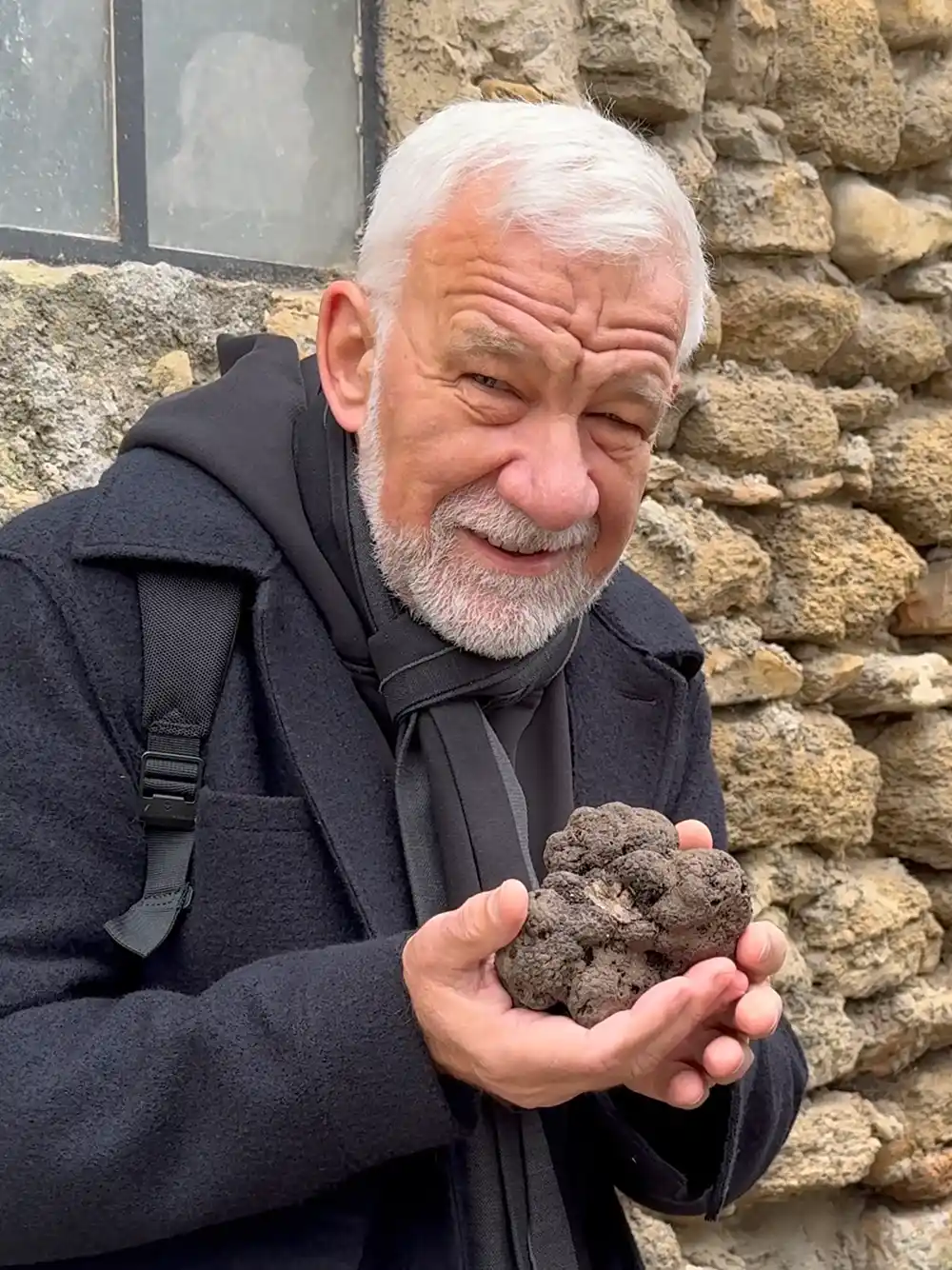 Trueffelmarkt-Richerenches-Trueffel Ein älterer Mann mit weißem Haar hält eine große schwarze Trüffel und lächelt vor einer Steinmauer.