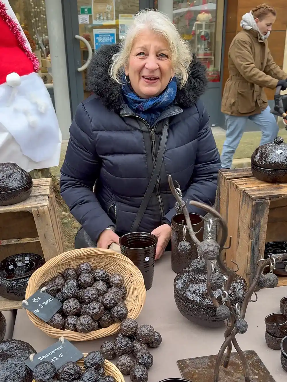 Trueffelmarkt-Richerenches-Trueffelmagnete Eine lächelnde Frau in einem dunklen Mantel steht an einem Marktstand im Freien mit Körben voller dunkler Backwaren und Töpferwaren.