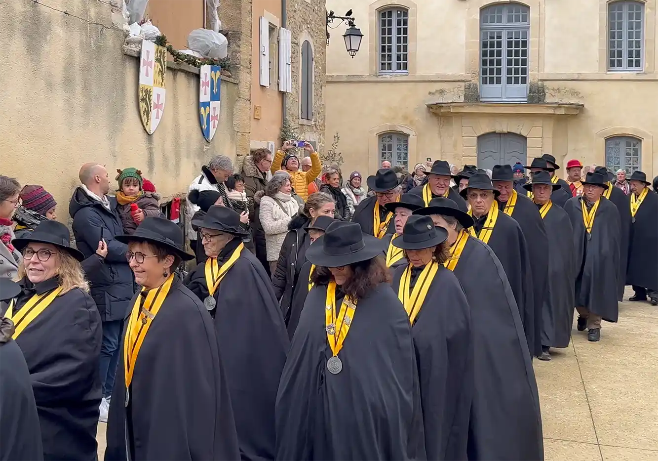 Trueffelmarkt-Richerenches-Umzug Menschen in schwarzen Umhängen und Hüten mit gelben Medaillen marschieren in einer Prozession, während Schaulustige zusehen und Fotos machen.