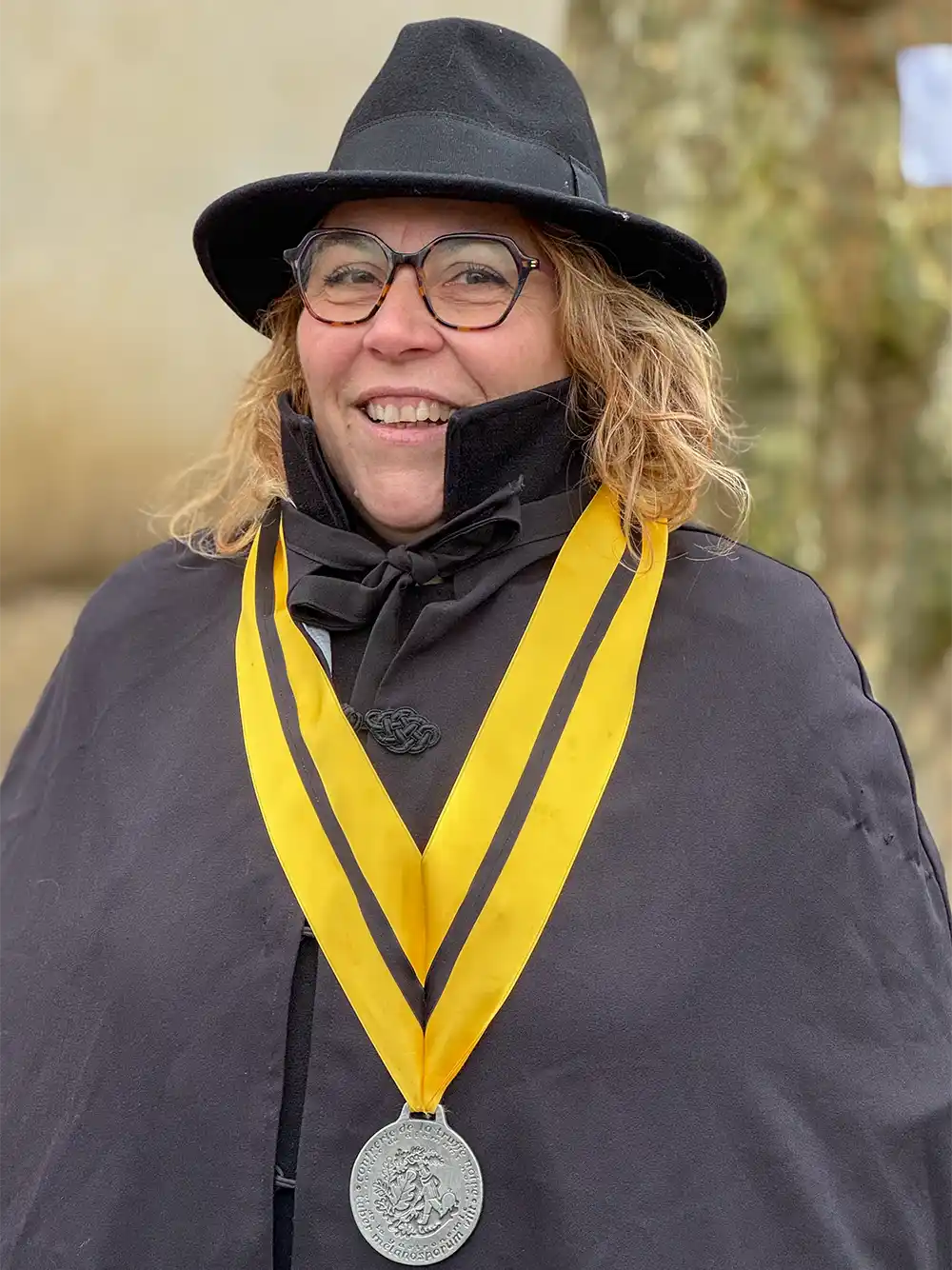 Trueffelmarkt-Richerenches-Virginie-Vincent-Jardin Eine lächelnde Person mit Brille und schwarzem Hut trägt einen Umhang und eine gelb gestreifte Medaille an einem Band um den Hals.