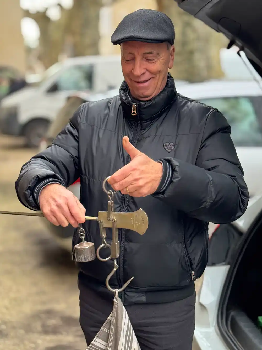 Trueffelmarkt-Richerenches-Waage Ein lächelnder Mann in schwarzer Jacke wiegt mit einer Handwaage einen Gegenstand im Freien neben einem geparkten Auto.