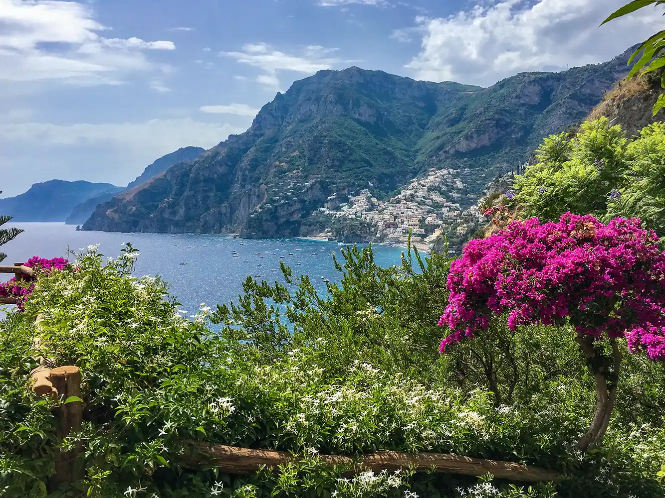 Amalfikueste-Positano Bunte Blumen mit Blick auf ein blaues Meer mit Bergen und einem Küstendorf in der Ferne unter einem teilweise bewölkten Himmel.