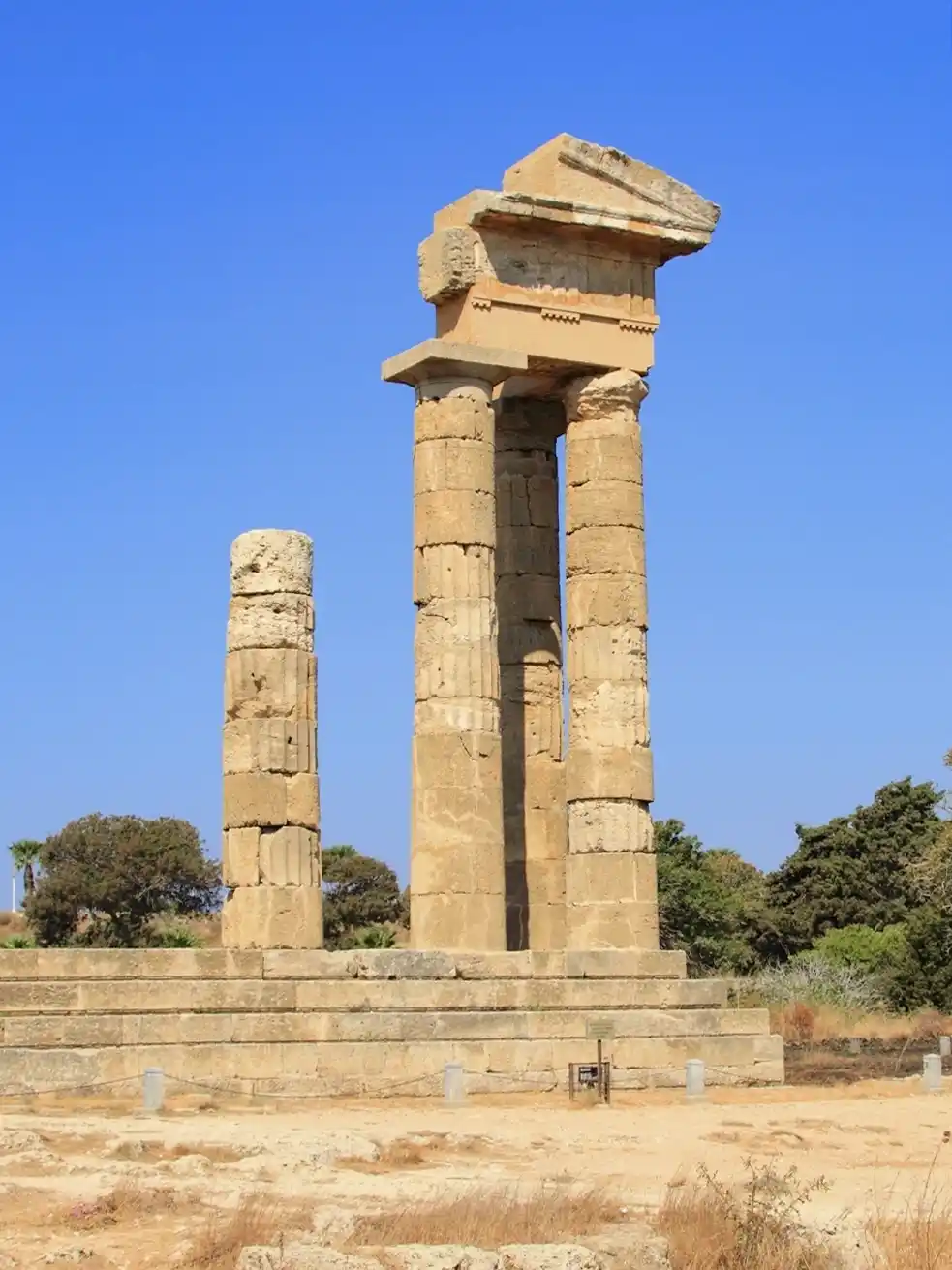 Die Ruinen der antiken Steinsäulen eines griechischen Tempels stehen vor einem strahlend blauen Himmel und Bäumen.