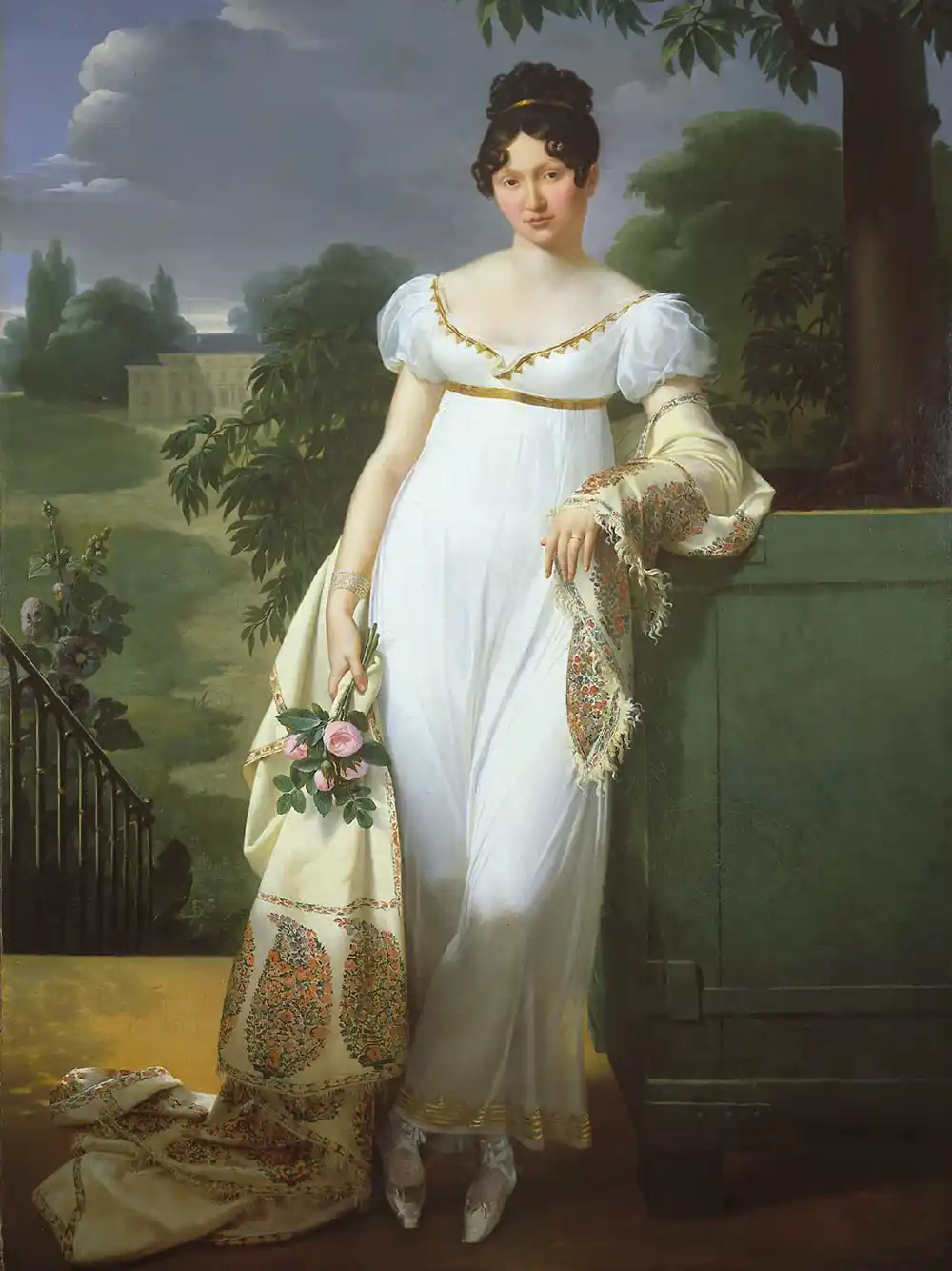 Eine Frau in einem weißen Regency-Kleid steht im Freien, hält einen Schal und Blumen in der Hand und ist von Grün umgeben.