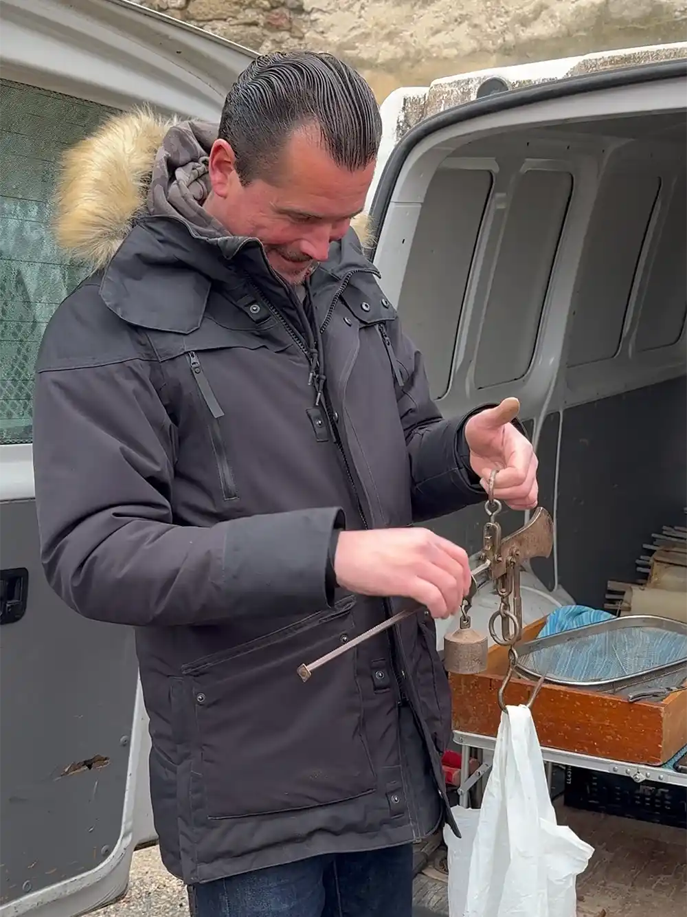 Ein Mann in einem schwarzen Wintermantel wiegt mit einer Hängewaage eine Plastiktüte in der Nähe einer offenen Lieferwagentür.