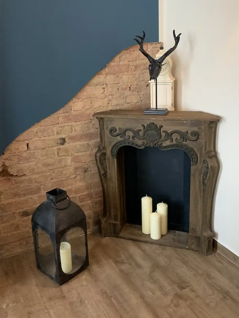 Dekorativer Kamin mit Kerzen, Laterne und Geweihskulptur vor einer blauen, freiliegenden Ziegelwand.